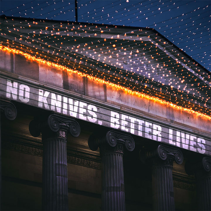 A retouched image of the same building in Glasgow at night, with No Knives Better Lives edited like a projection onto the building.
